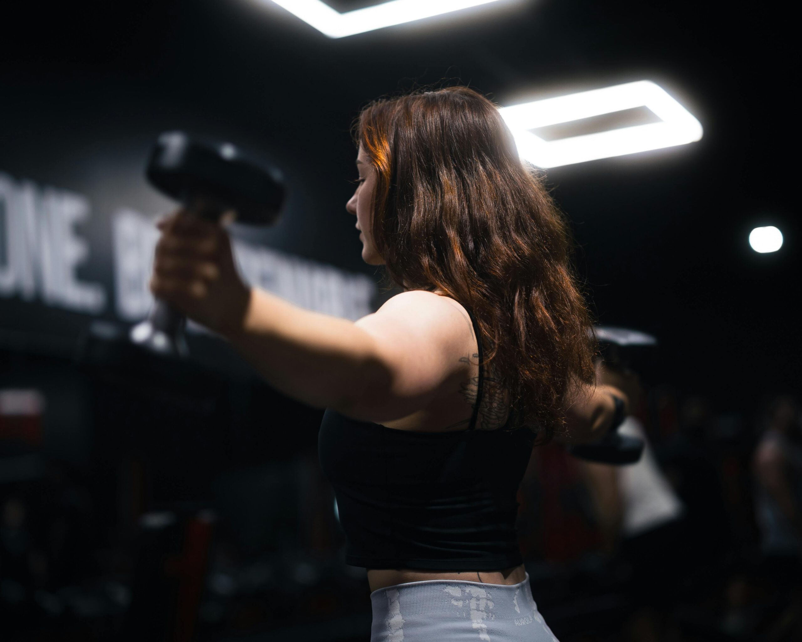 Side view of a woman exercising with dumbbells in a gym under bright lighting.